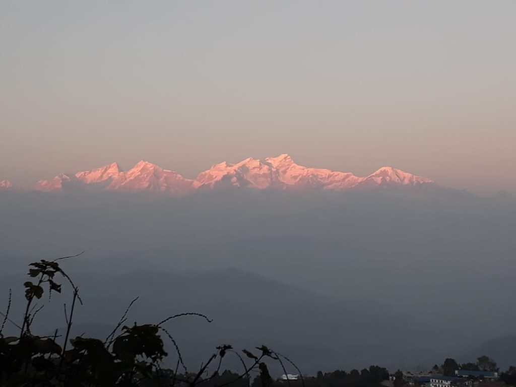 A photograph capturing the breathtaking mountain scenery from Chisapani during sunset, with warm hues painting the sky and mountains in a serene evening light.