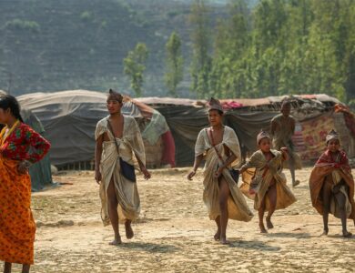 Raute family around their camp in a forest clearing, with traditional shelters in the background.