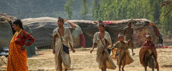 Raute family around their camp in a forest clearing, with traditional shelters in the background.