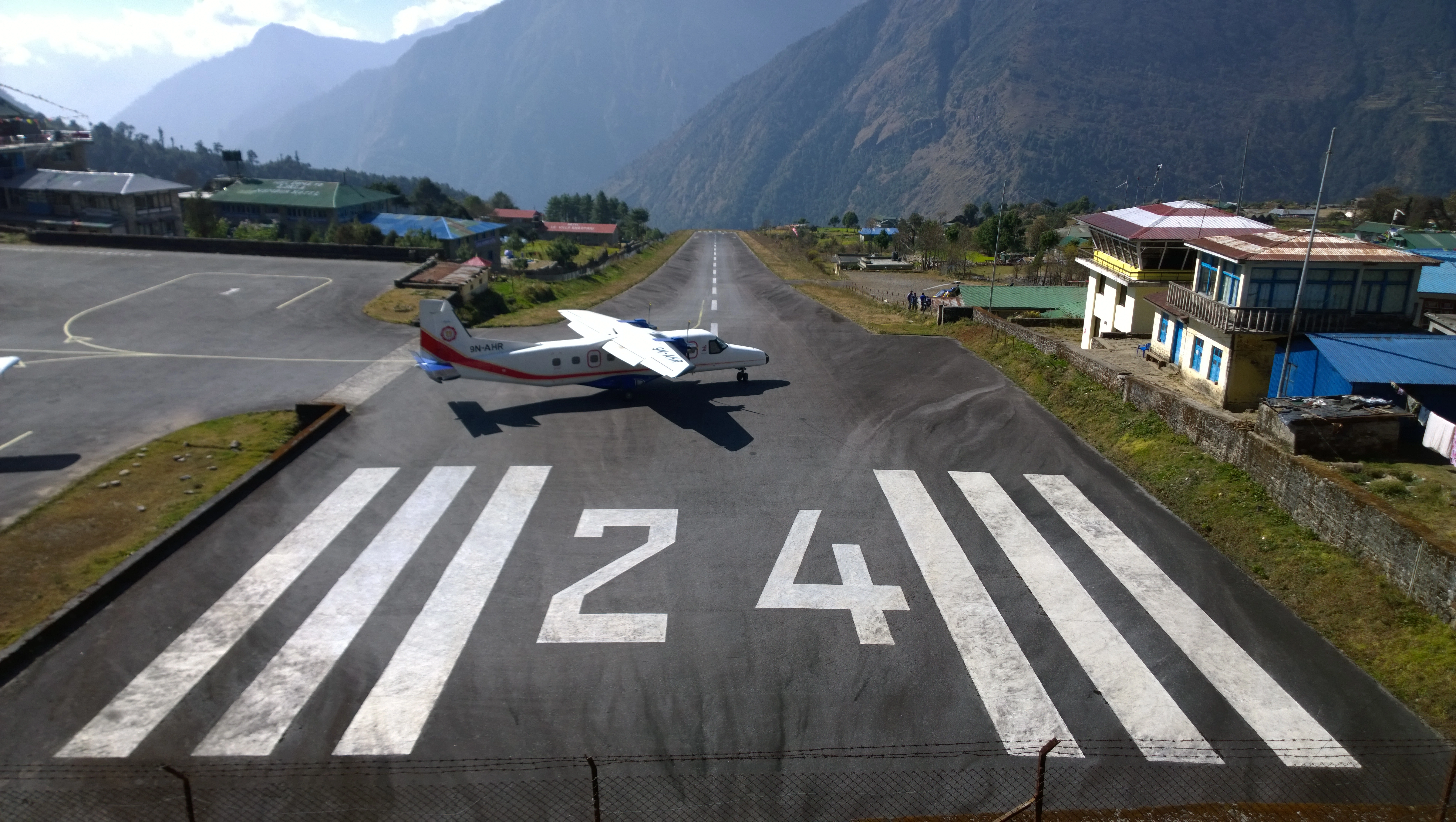 An aerial view of Lukla Airport, nestled in the mountains, with a small plane on the runway ready for takeoff.