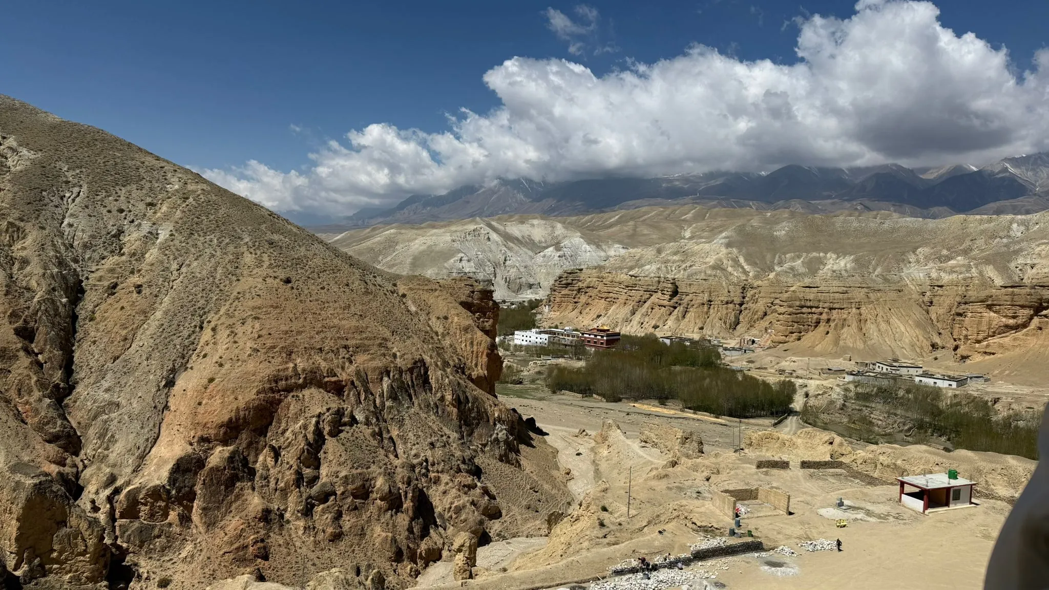 Panoramic landscape view of Upper Mustang, Nepal, featuring dry desert hills, layered rock cliffs, scattered settlements, and dramatic Himalayan mountains beneath large white clouds.