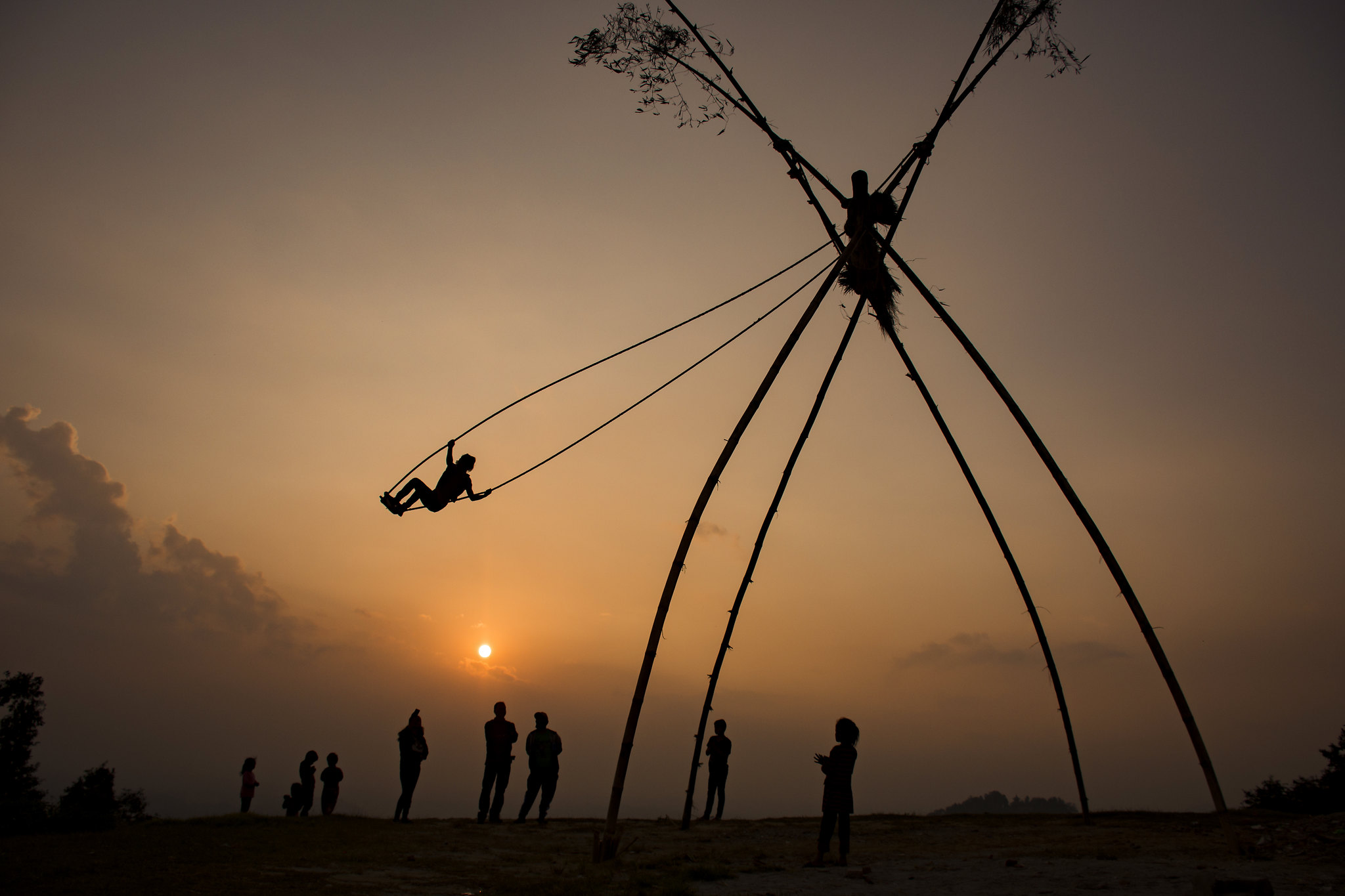A traditional Nepali bamboo swing (ping) set up in a rural area during the Dashain and Tihar festival, with children joyfully swinging high. The swing represents spiritual cleansing and the excitement of the festive season, as villagers gather to celebrate."