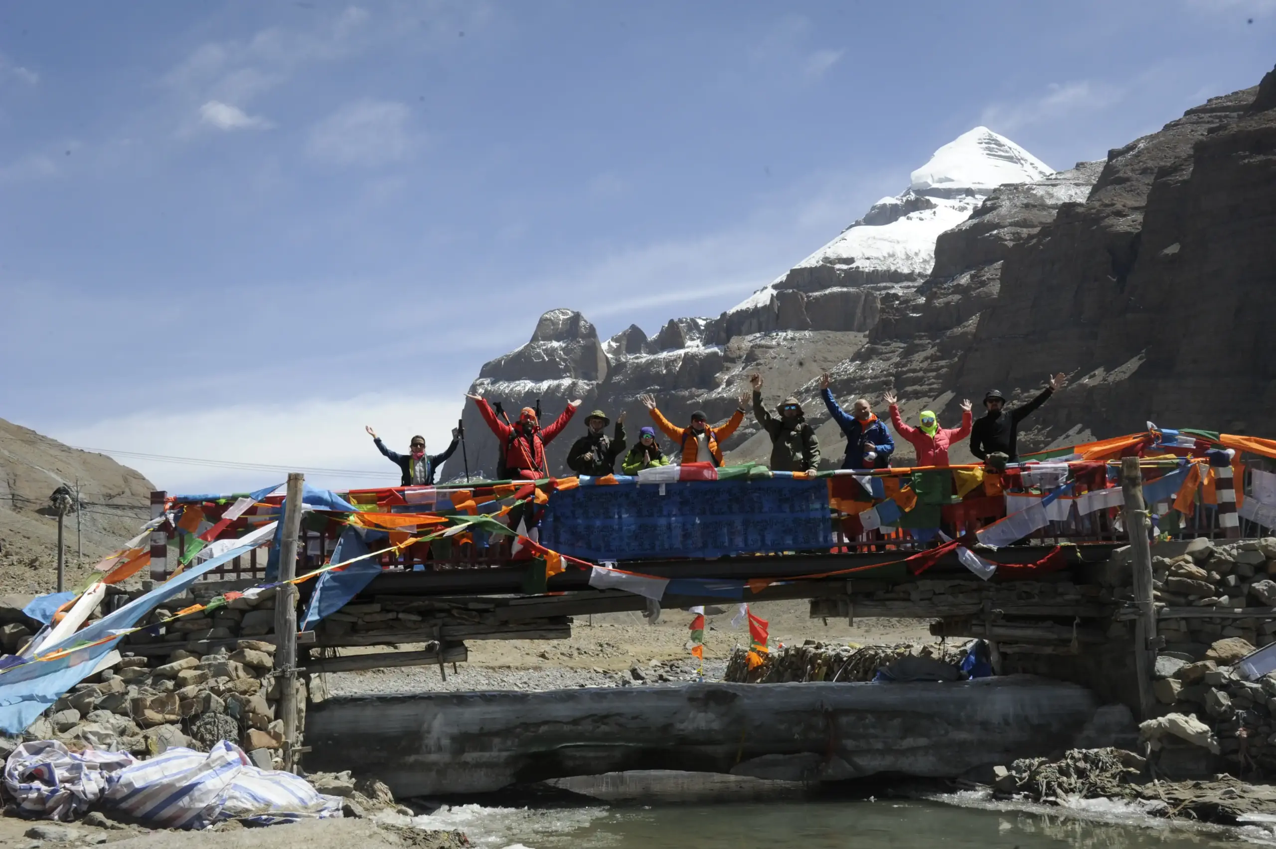 Pilgrimage group celebrating on a prayer flag bridge with Mount Kailash during Kailash Mansarovar Yatra 2026