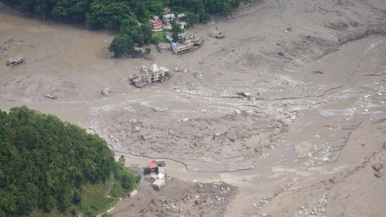 Aerial view of severe flood and debris damage in a Himalayan valley, with houses and roads surrounded by mud, rocks, and floodwater.
